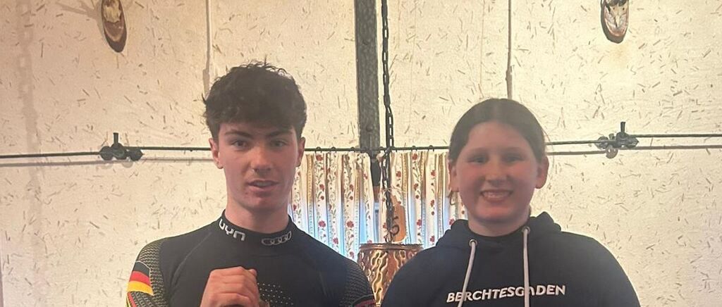 Zwei Personen, ein Mann und eine Frau, stehen nebeneinander in einem Raum. Der Mann trägt einen schwarzen Neoprenanzug mit Logo, und die Frau trägt eine schwarze Kapuzenjacke mit weißem Text. Hinter ihnen befindet sich eine Wand mit einem weißen Vorhang mit roten Blumen und einem hängenden Objekt.