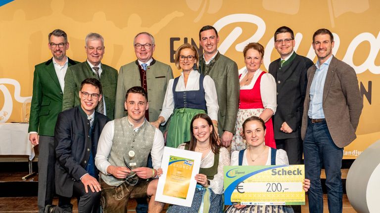 Eine Gruppe von Menschen in traditioneller deutscher Kleidung posiert für ein Foto. Zwei Frauen halten ein Zertifikat und einen Scheck. Der Mann in der Mitte hält eine Medaille. Im Hintergrund befinden sich die Buchstaben 'E' und 'R'.