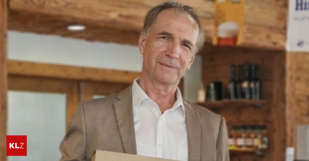 Ein älterer Mann in einem braunen Anzug hält eine Box, lächelt und steht in einem Raum mit Holzwänden und Regalen.