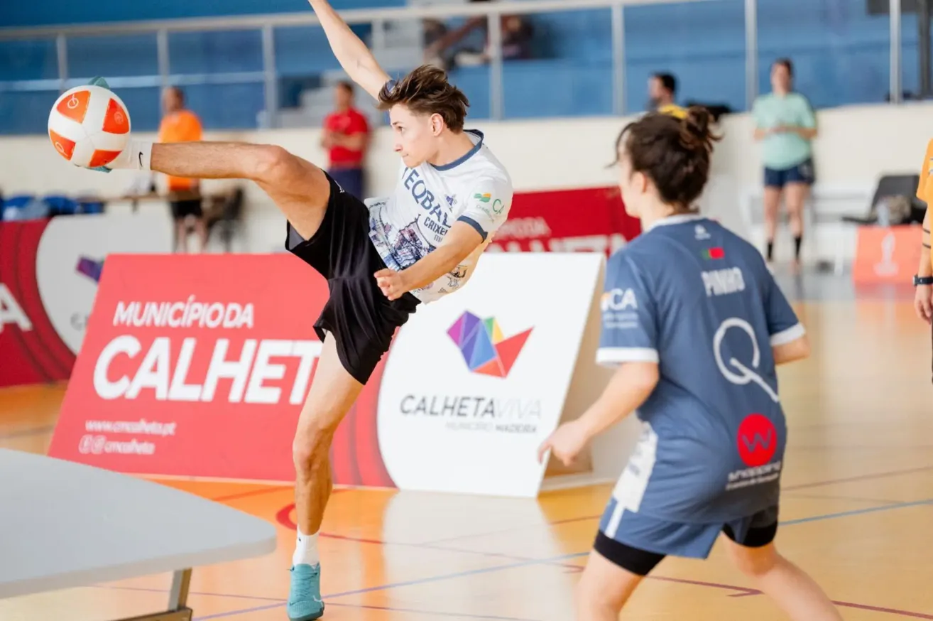 Ein junger Athlet tritt einen Ball in einer Turnhalle, trägt ein Trikot mit dem Wort 'Cricket' darauf. Hinter ihm ist ein Banner mit einem bunten Logo zu sehen, und ein anderer Spieler ist in einem blauen Trikot.