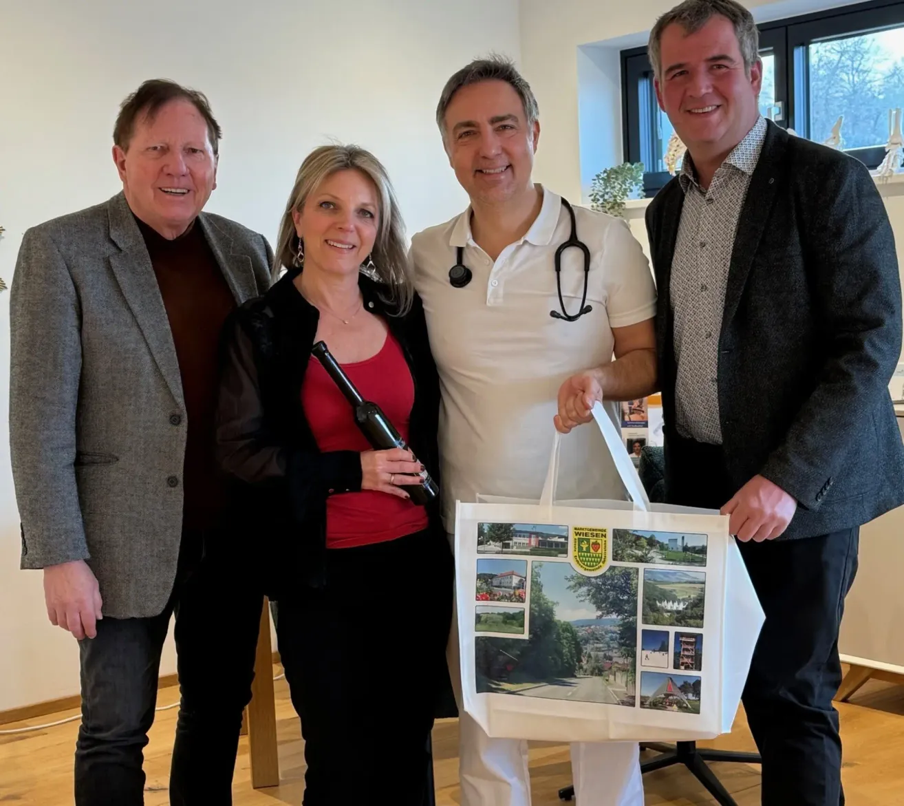 Vier Erwachsene stehen lächelnd zusammen. Ein Mann in einem weißen Hemd mit Stethoskop hält eine weiße Tasche mit Bildern. Die Frau neben ihm hält eine Flasche. Dahinter ist eine Wand mit einer Pflanze.