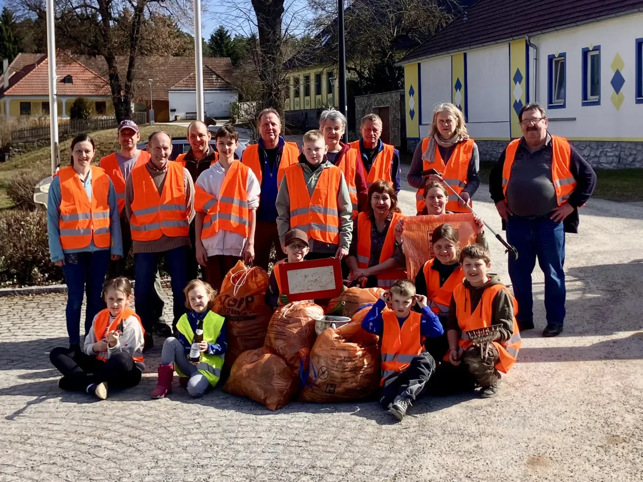 Eine Gruppe von Menschen in orangefarbenen Westen posiert für ein Foto mit Müllsäcken vor einem Haus und einem Baum.