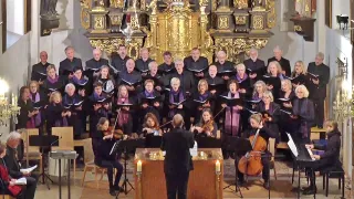 Ein Chor steht in einer Kirche mit Violinen und Celli. Der Dirigent leitet vom Notenständer aus, mit Kerzen in der Nähe. Die Chormitglieder singen und halten Bücher.