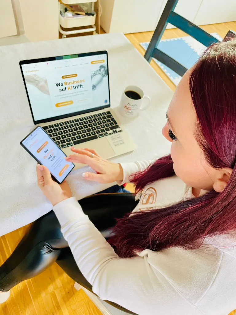 Eine Frau mit roten Haaren sitzt an einem Tisch und schaut auf ihr Handy. Ein Laptop mit einer Website auf dem Bildschirm steht auf dem Tisch. Ein Kaffeebecher steht neben dem Laptop.