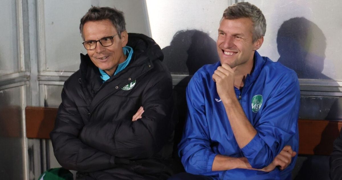 Zwei Männer, einer in einer schwarzen Jacke und der andere in einer blauen Jacke, lächeln und posieren für ein Foto mit verschränkten Armen.