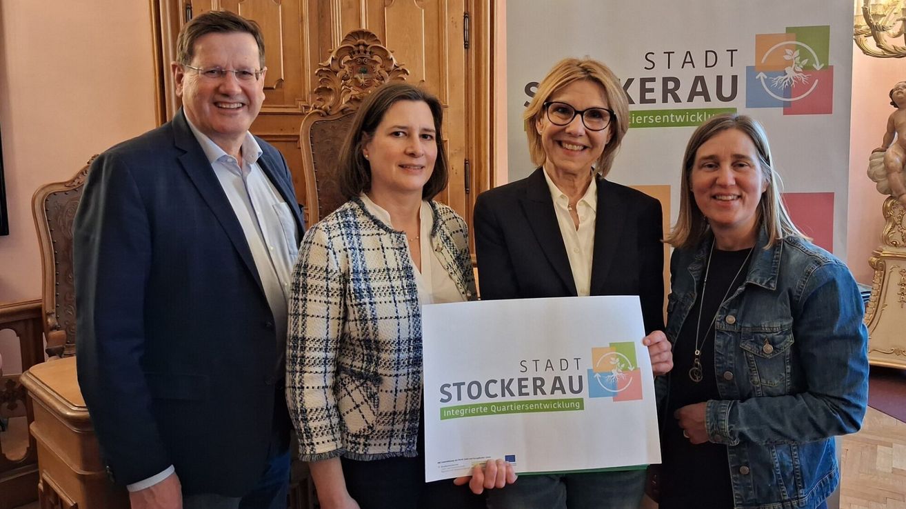 Vier Menschen lächeln für ein Foto vor einer Holzwand mit einem Banner. Zwei Frauen halten ein Plakat mit 'Stadt Stockerau' darauf. Der Mann und eine weitere Frau lächeln.