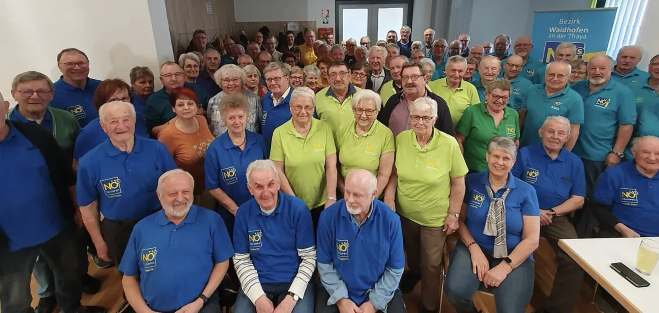 Eine Gruppe älterer Personen, sowohl Männer als auch Frauen, versammelt sich in einem Raum und trägt blaue und grüne Poloshirts. Sie scheinen für ein Foto zu posieren.