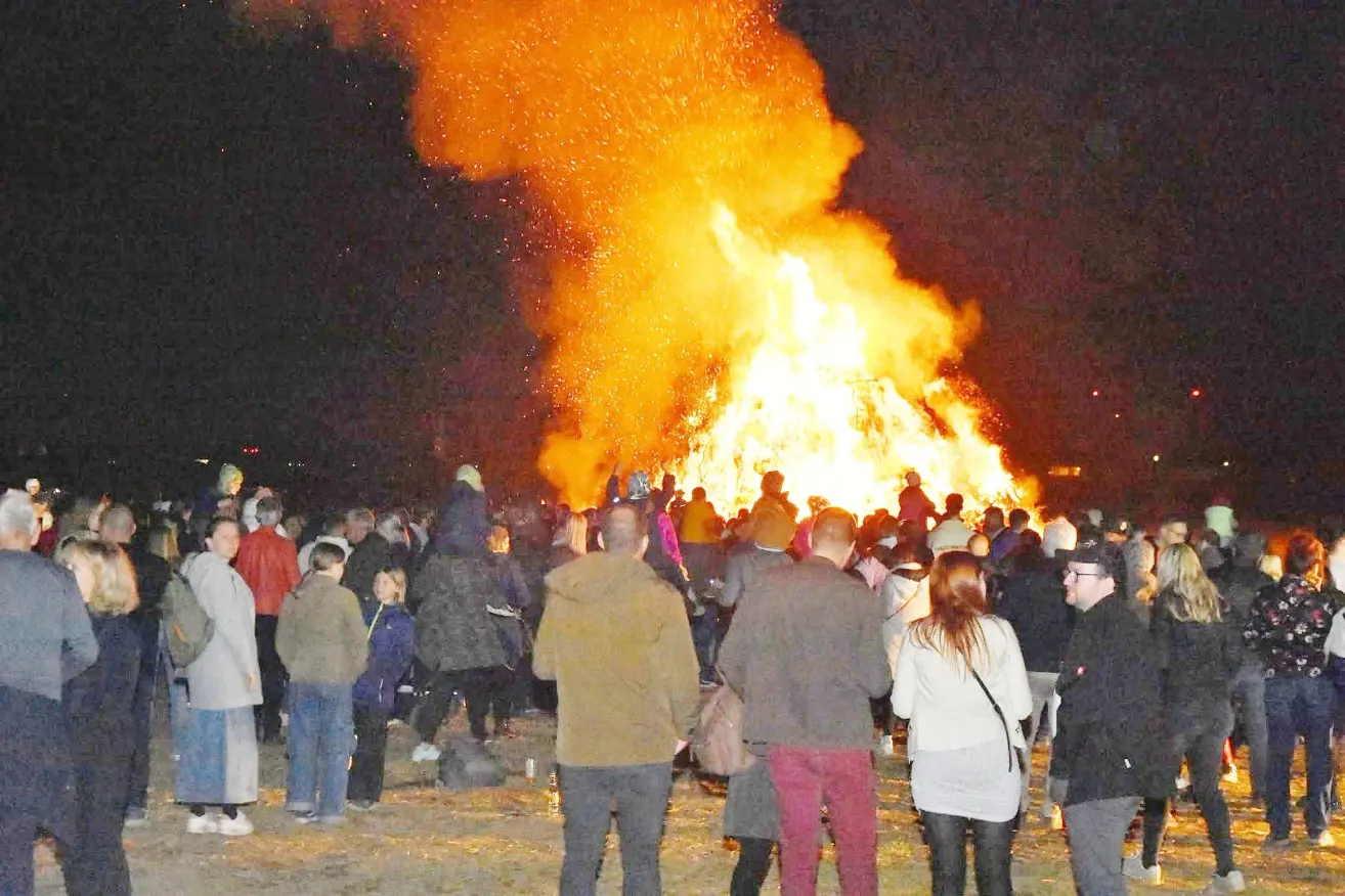Ein großes Lagerfeuer brennt bei Nacht hell und wird von einer Menschenmenge umringt. Einige stehen dicht am Feuer, andere weiter hinten. Viele tragen Hüte und Jacken.