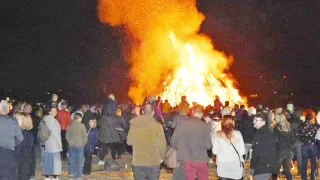 Ein großes Lagerfeuer brennt bei Nacht hell und wird von einer Menschenmenge umringt. Einige stehen dicht am Feuer, andere weiter hinten. Viele tragen Hüte und Jacken.