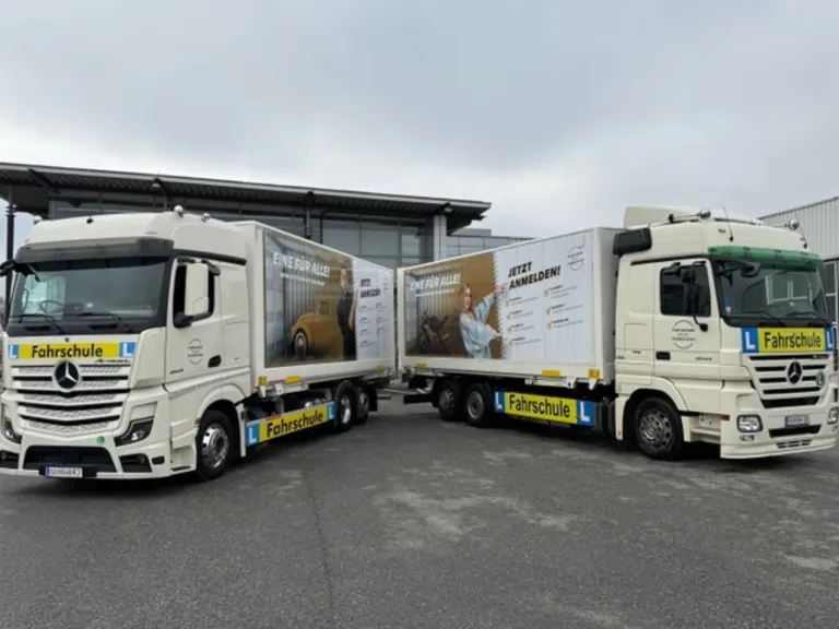Zwei weiße LKWs sind auf einem Parkplatz geparkt. Ein LKW hat eine Werbung für eine Fahrschule.
