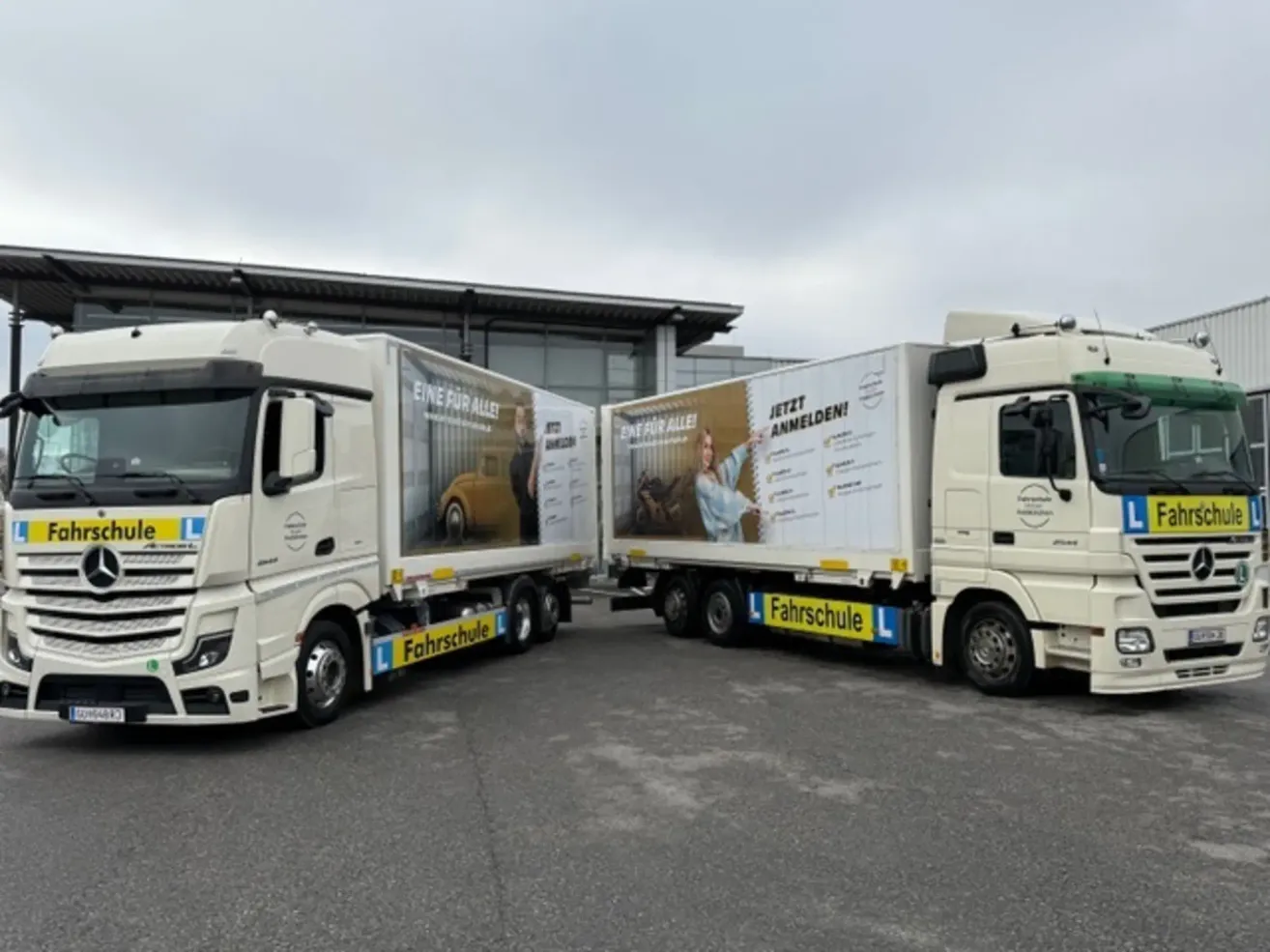 Zwei weiße LKWs sind auf einem Parkplatz geparkt. Ein LKW hat eine Werbung für eine Fahrschule.