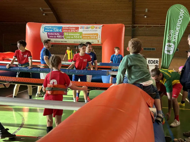 Eine Gruppe junger Kinder und Erwachsener spielt in einem Hüpfhaus, wobei ein Erwachsener hinter ihnen steht. Sie sind in einer Sporthalle mit einem Banner an der Wand.