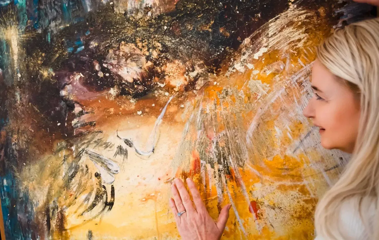 Ein abstraktes Gemälde mit einer Mischung aus schwarzen, braunen und gelben Farbtönen. Eine Hand berührt das Gemälde, geschmückt mit einem Ring. Das Gemälde hat eine strukturierte, vielschichtige Optik.