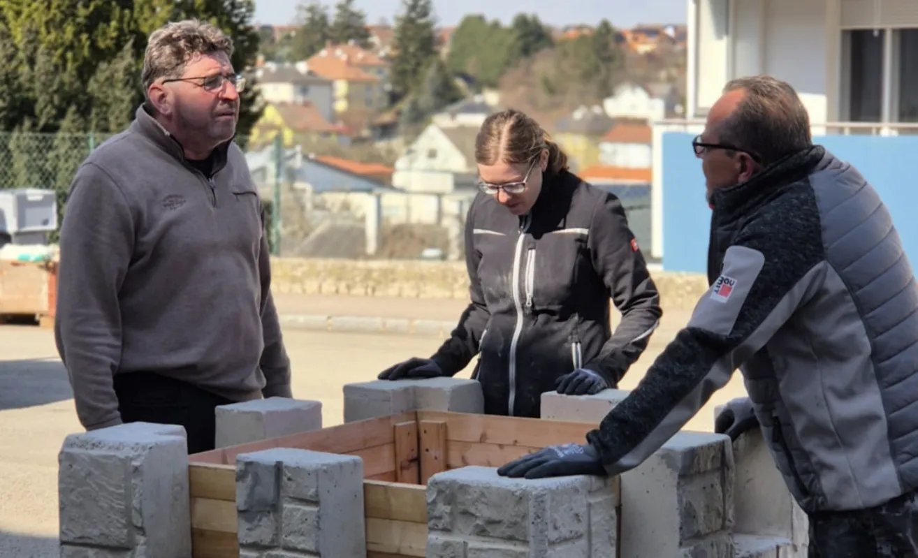 Drei Personen in Winterkleidung stehen um eine hölzerne Kastenstruktur herum. Die Frau trägt eine Brille und eine schwarze Jacke. Der Mann zu ihrer Linken trägt eine graue Jacke. Der Mann zu ihrer Rechten trägt eine graue Jacke mit weißen Streifen.