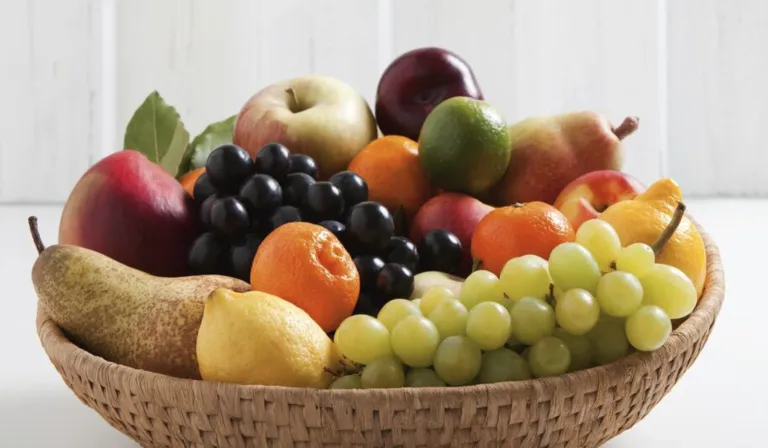 Ein Korb aus Weide gefüllt mit verschiedenen Früchten: Äpfel, Pflaumen, Orangen, Trauben und eine Limette. Die Früchte sind so angeordnet, dass ihre Farben und Texturen zur Geltung kommen.