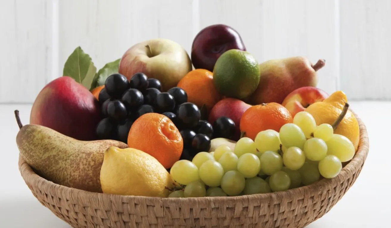 Ein Korb aus Weide gefüllt mit verschiedenen Früchten: Äpfel, Pflaumen, Orangen, Trauben und eine Limette. Die Früchte sind so angeordnet, dass ihre Farben und Texturen zur Geltung kommen.