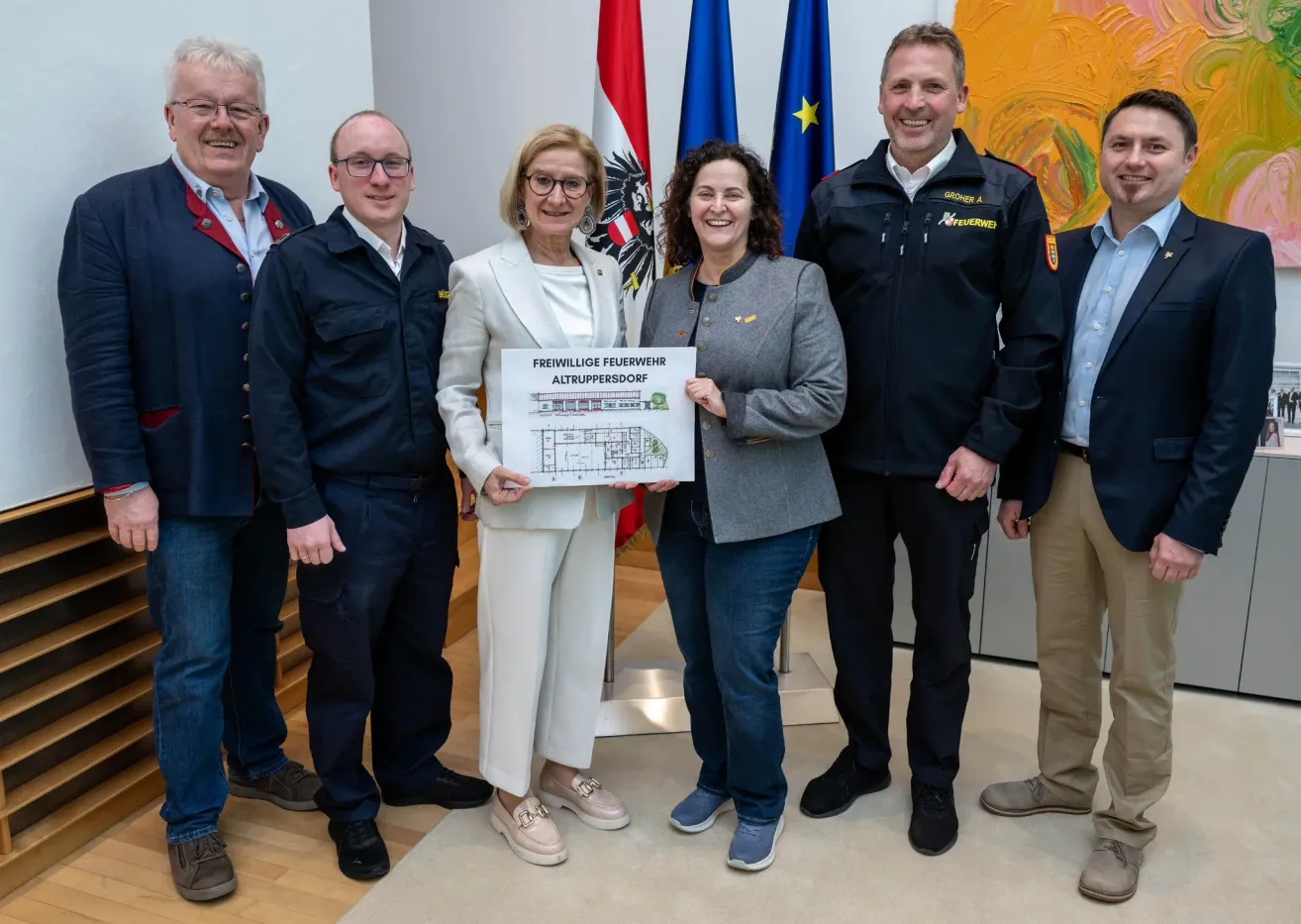 Fünf Personen stehen in einem Raum und lächeln. Zwei Frauen halten ein Papier mit Zeichnungen. Hinter ihnen befinden sich Fahnen. Ein Mann trägt eine Medaille.