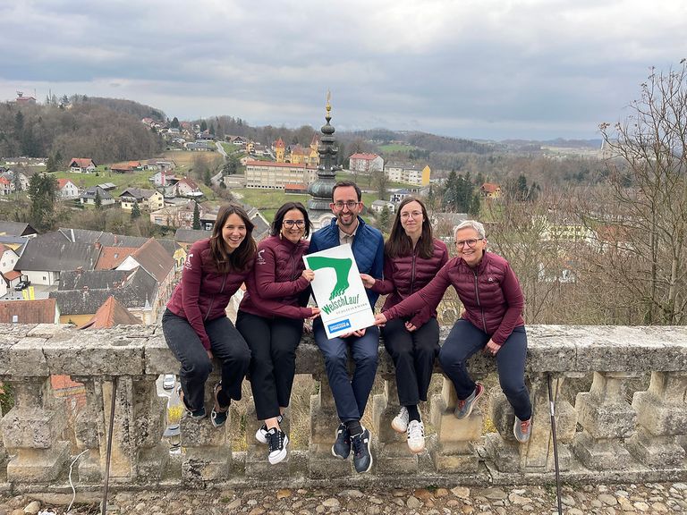 Fünf Personen posieren für ein Foto auf einer Steinmauer mit einer malerischen Aussicht dahinter. Sie tragen alle weinrote Jacken. Der Mann in der Mitte hält ein grünes Schild.