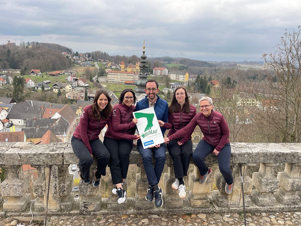 Fünf Personen posieren für ein Foto auf einer Steinmauer mit einer malerischen Aussicht dahinter. Sie tragen alle weinrote Jacken. Der Mann in der Mitte hält ein grünes Schild.