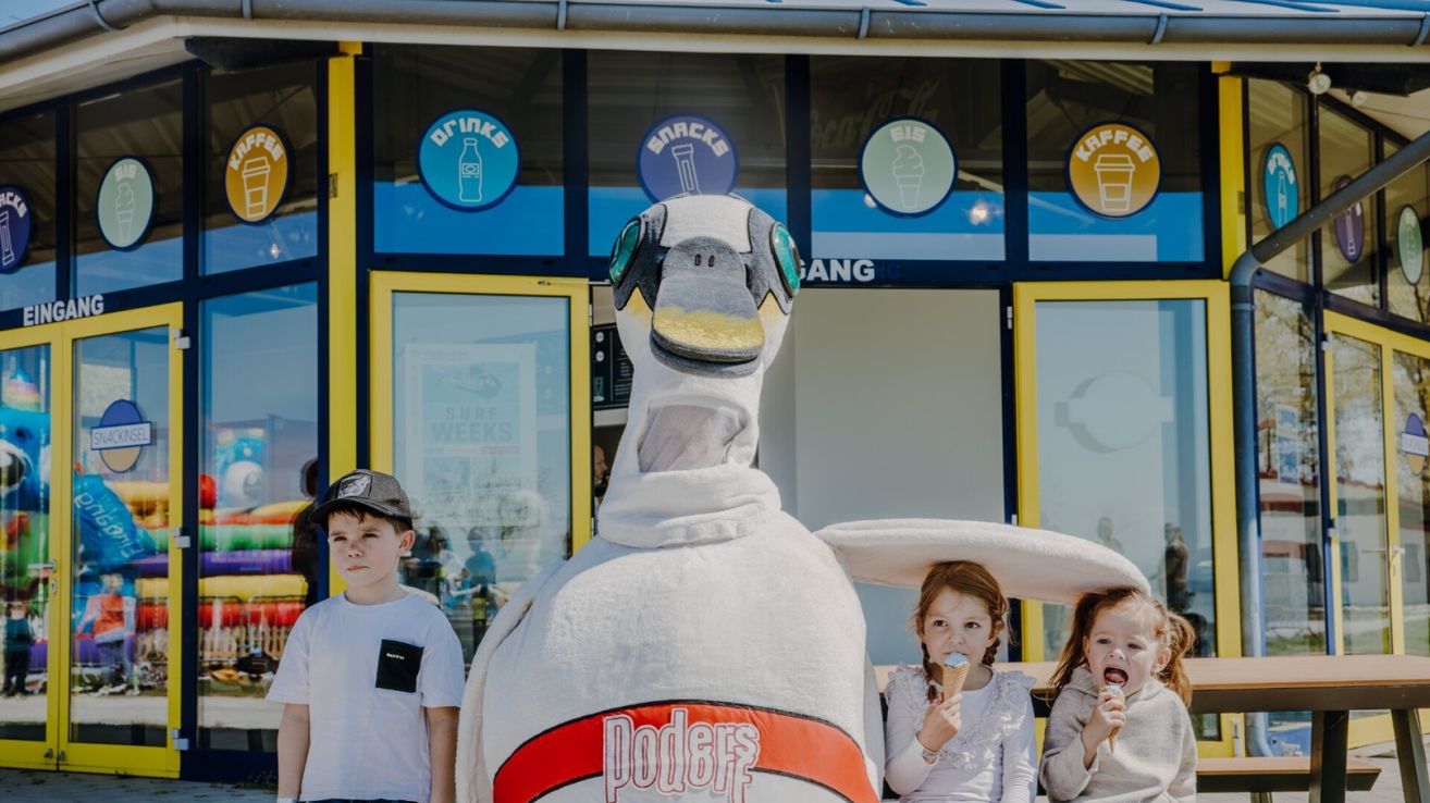 Drei Kinder und eine Pinguin-Maskottchen sind vor einem Geschäft. Das Geschäft hat Schilder für Getränke, Snacks, Eis und Kaffee. Die Kinder essen Eis, und der Pinguin trägt ein Kostüm.
