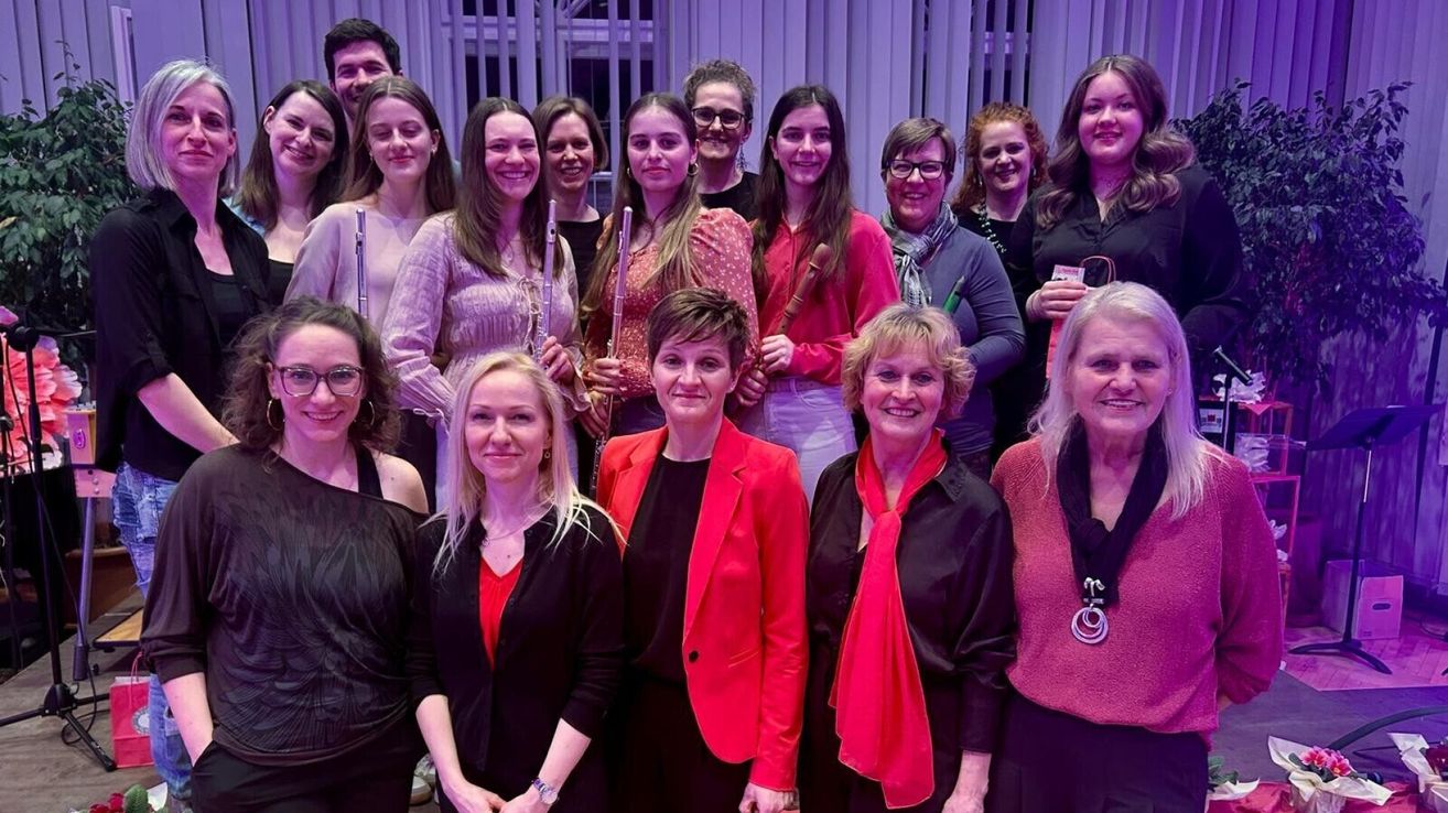 Eine Gruppe von Frauen, einige halten Musikinstrumente, lächeln für ein Gruppenfoto unter lila Beleuchtung. Sie tragen verschiedene Kleidung, einschließlich Kleider, Schals und Blazer.