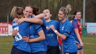 Vier junge Frauen in blauen Trikots, eines mit Logo, feiern. Sie umarmen sich gegenseitig in einem Park mit verschwommenen Bäumen im Hintergrund.
