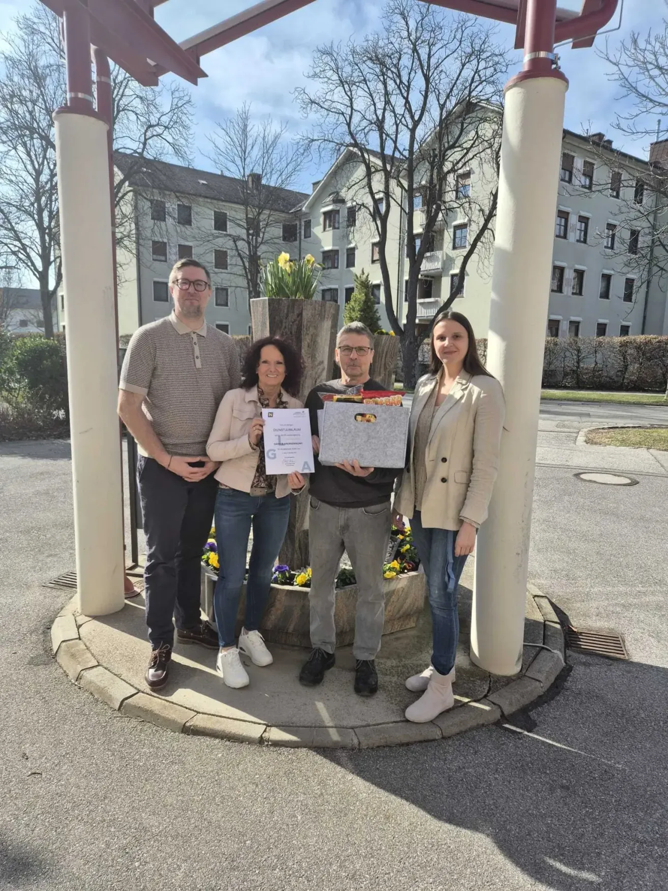 Vier Personen stehen auf einem Kreisverkehr mit Gebäuden im Hintergrund. Ein Mann hält eine Box und ein Zertifikat, während die anderen Zertifikate halten.