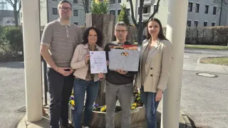 Vier Personen stehen auf einem Kreisverkehr mit Gebäuden im Hintergrund. Ein Mann hält eine Box und ein Zertifikat, während die anderen Zertifikate halten.