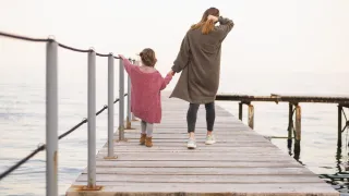 Eine Frau und ein junges Mädchen gehen gemeinsam auf einem hölzernen Steg über das Wasser. Das Mädchen hält die Hand der Frau und die Frau sieht nachdenklich aus.