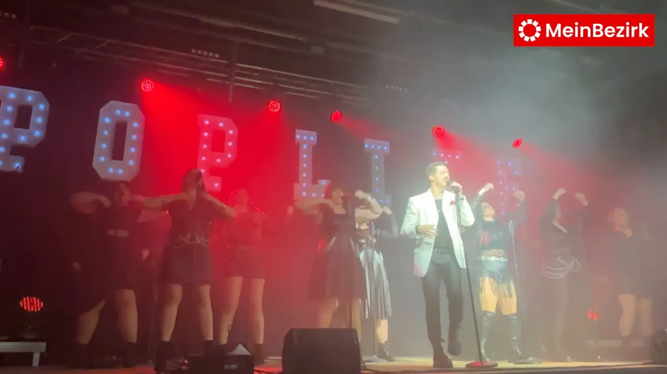 Ein Künstler singt auf der Bühne mit einem Mikrofon. Hinter ihm tanzen mehrere Frauen in schwarzen Kleidern. Die Bühne ist mit roten Lichtern beleuchtet und große beleuchtete Buchstaben buchstabieren das Wort 'Pili.'