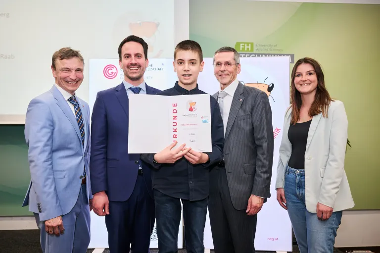 Fünf Erwachsene und ein junger Junge in Anzügen stehen hinter einem Banner. Der Junge hält ein Zertifikat und lächelt. Zwei Männer an den Seiten tragen Krawatten.