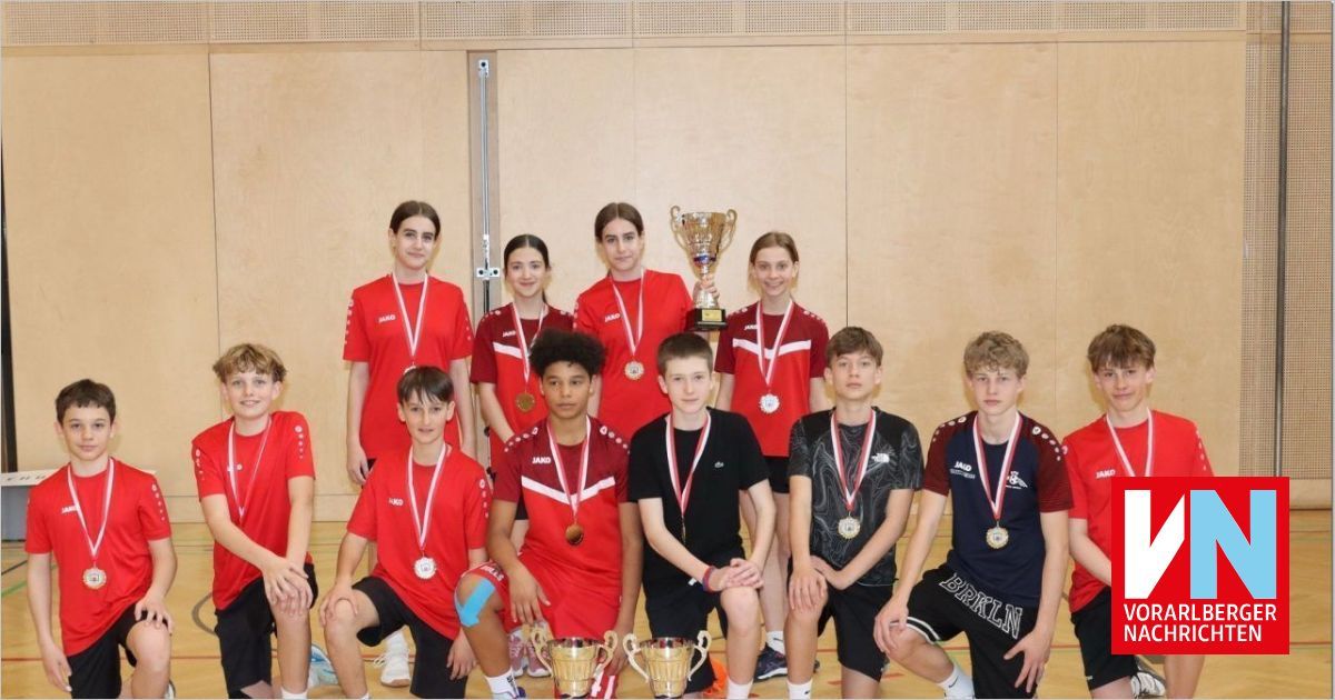 Eine Gruppe junger Athleten, die rote Trikots und Medaillen tragen, posiert für ein Foto in einer Turnhalle. Sie halten eine Trophäe und einige knien.