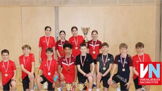 Eine Gruppe junger Athleten, die rote Trikots und Medaillen tragen, posiert für ein Foto in einer Turnhalle. Sie halten eine Trophäe und einige knien.