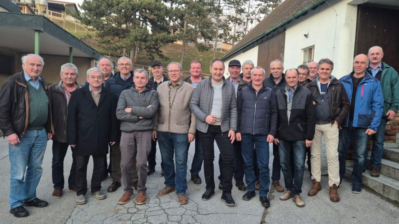 Eine Gruppe von Männern steht zusammen draußen. Sie alle lächeln und scheinen für ein Foto zu posieren. Sie tragen Winterkleidung und einige von ihnen haben Brillen. Hinter ihnen sind Bäume und ein Gebäude.