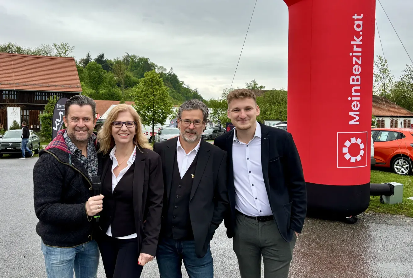 Vier Personen stehen vor einem roten aufblasbaren Objekt. Sie tragen formelle Kleidung. Dahinter sind Autos geparkt. Im Hintergrund befinden sich Bäume und Häuser.