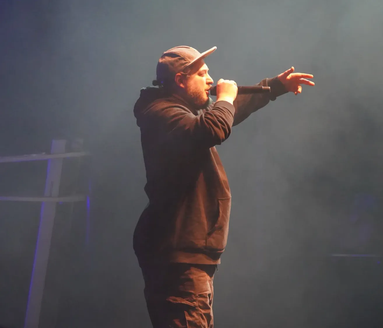 Ein Mann steht auf der Bühne, singt in ein Mikrofon, mit ausgestreckten Armen, trägt eine Mütze und eine Kapuzenjacke.