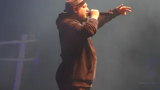 Ein Mann steht auf der Bühne, singt in ein Mikrofon, mit ausgestreckten Armen, trägt eine Mütze und eine Kapuzenjacke.