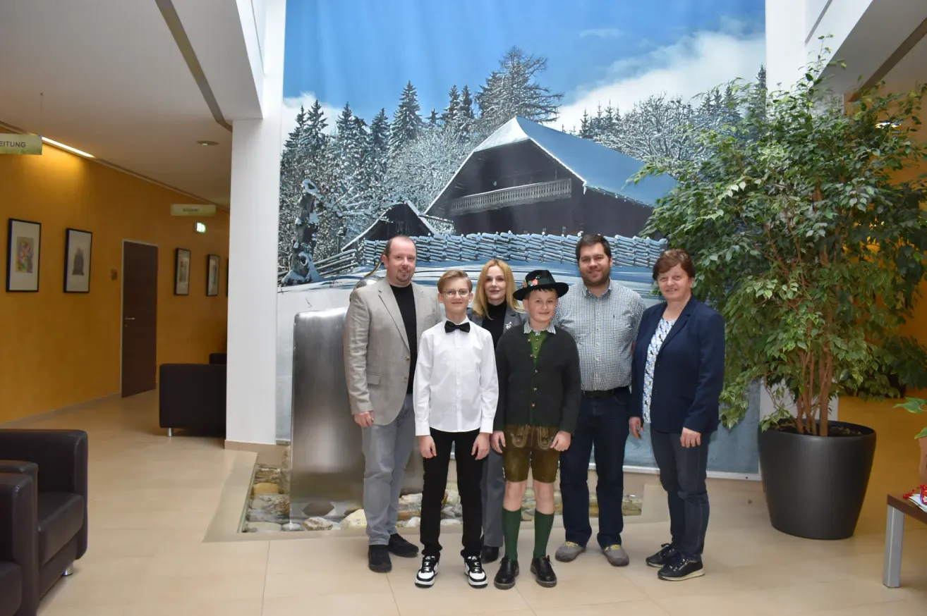 Eine Familie steht vor einer Winterszene-Wandmalerei, wobei zwei Jungen traditionelle Kleidung tragen und Erwachsene formell gekleidet sind. Die Umgebung scheint drinnen zu sein, mit einem Brunnen und einer Pflanze rechts.