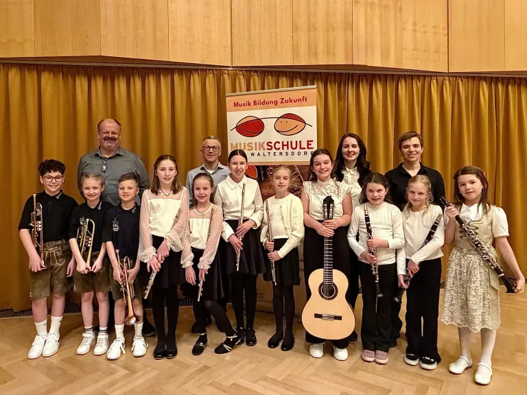 Eine Gruppe von Kindern und Erwachsenen posiert für ein Foto. Sie halten Musikinstrumente. Hinter ihnen steht ein Banner mit der Aufschrift 'Musikschule Waltersdorf'. Der Ort scheint eine Musikschule zu sein.