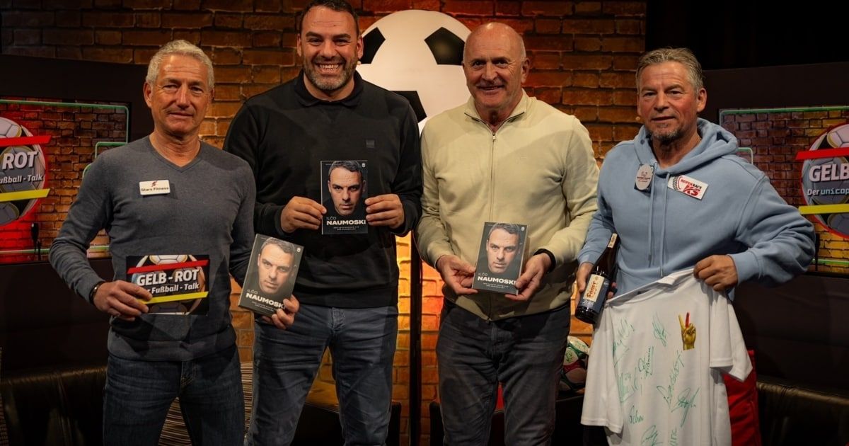 Vier Männer stehen eng beieinander, halten Bücher und eine Flasche, vor einem Fußball-Hintergrund, tragen Freizeitkleidung und lächeln.