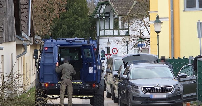 Ein Mann steht neben einem gepanzerten Fahrzeug auf einer Straße mit Geschwindigkeitsbegrenzungsschildern. Mehrere Autos sind am Straßenrand geparkt, darunter ein grauer SUV mit offenem Kofferraum.