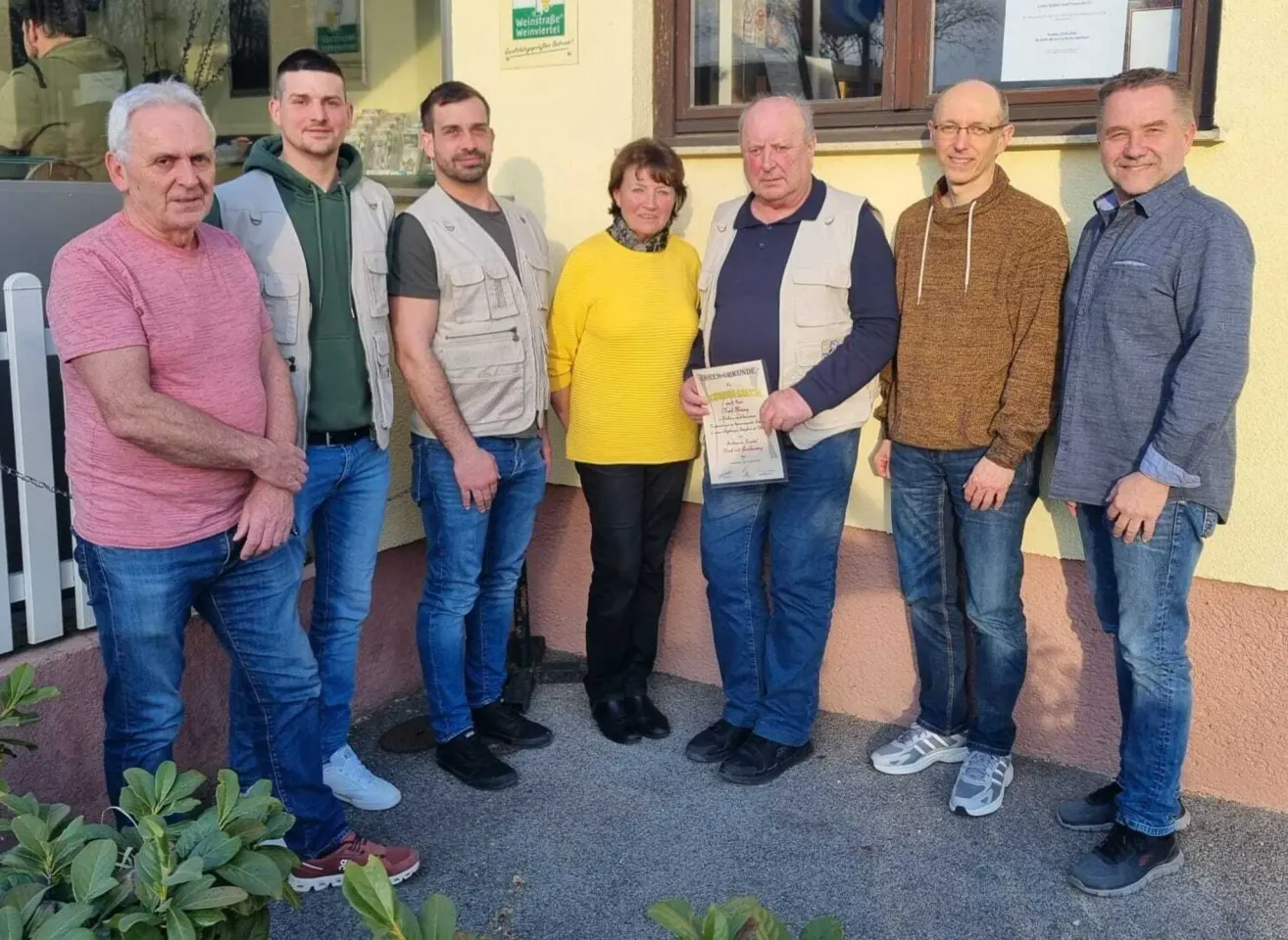 Eine Gruppe von sechs Erwachsenen steht vor einem Gebäude und lächelt für ein Foto. Eine Frau in der Mitte hält ein Zertifikat. Sie tragen Freizeitkleidung, und ein Schild ist an der Wand hinter ihnen sichtbar.