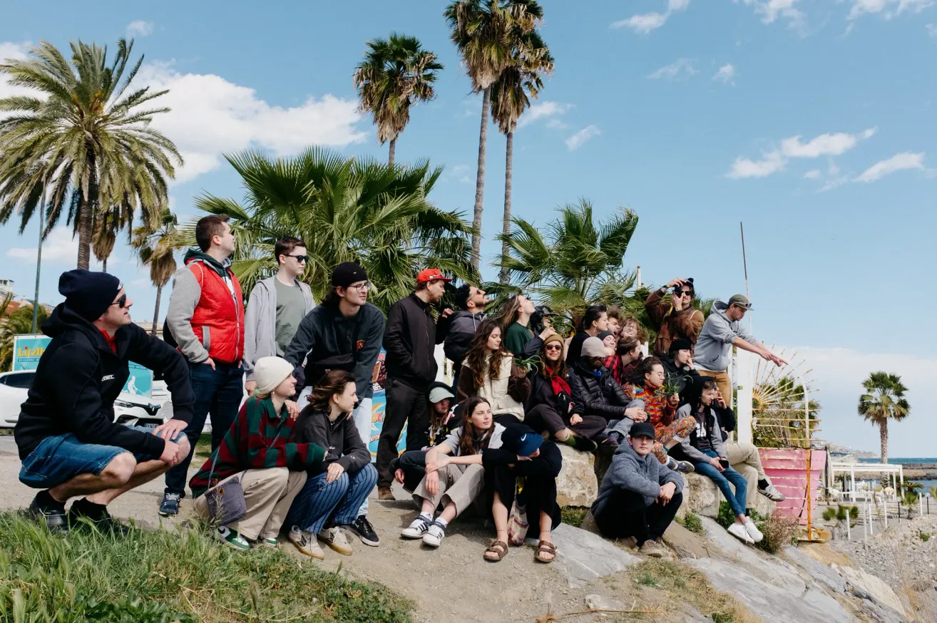 Eine Gruppe von Menschen versammelt sich auf einem Hügel mit Palmen. Einige sitzen auf dem Boden, während andere stehen. Die Atmosphäre ist entspannt, mit Personen in lässiger Kleidung. Der Himmel ist blau mit verstreuten Wolken.