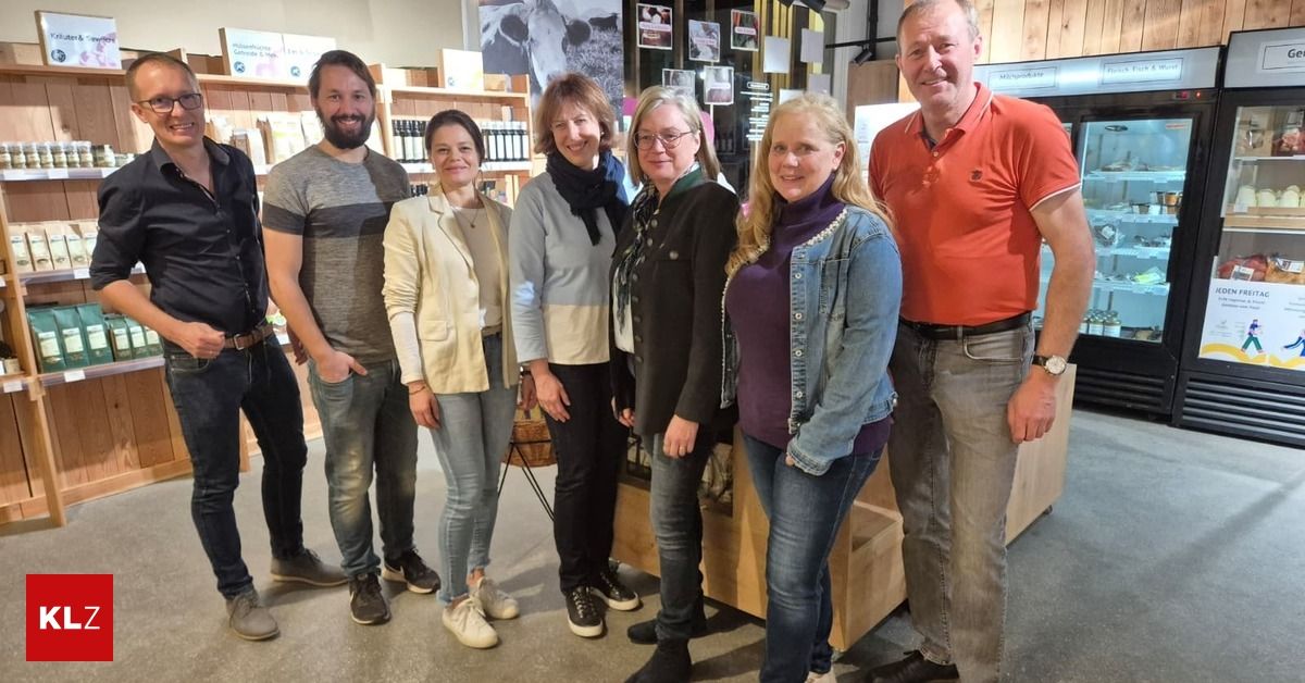 Eine Gruppe von Menschen steht in einem Geschäft, lächelt und posiert für ein Foto. Sie sind lässig gekleidet, einige tragen Brillen und andere halten Gegenstände. Ein Holzregal und ein Kuhposter sind im Hintergrund zu sehen.