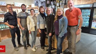 Eine Gruppe von Menschen steht in einem Geschäft, lächelt und posiert für ein Foto. Sie sind lässig gekleidet, einige tragen Brillen und andere halten Gegenstände. Ein Holzregal und ein Kuhposter sind im Hintergrund zu sehen.