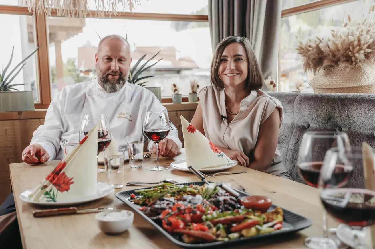 Ein Koch und eine Frau sitzen lächelnd an einem Tisch in einem Restaurant. Der Tisch ist mit Weingläsern, Tellern und einer großen Gemüseplatte gedeckt. Hinter ihnen befindet sich ein Fenster mit Blick auf ein Gebäude.