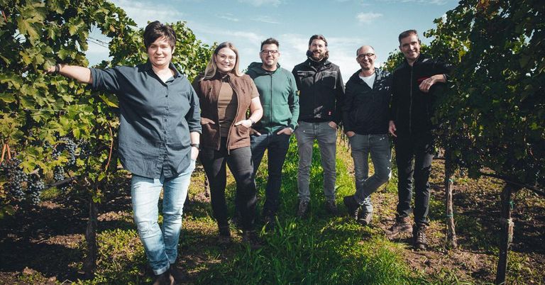 Eine Gruppe von sechs Personen, bestehend aus drei Frauen und drei Männern, steht zusammen in einem Weinberg. Sie alle lächeln und posieren für ein Foto.