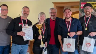 Vier Personen posieren für ein Foto mit Medaillen und Trophäen; eine Frau hält eine goldene Trophäe und ein Mann rechts hält eine weiße Tasche.