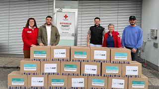 Fünf Personen stehen lächelnd hinter einem Stapel Kartons, die mit Illustrationen und den Worten 'Kumanya Kampanyas' beschriftet sind. Über ihnen steht ein Schild mit der Aufschrift 'Rotkreuz Mark'. Die Umgebung ist im Freien.