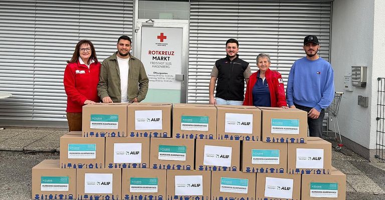 Fünf Personen stehen lächelnd hinter einem Stapel Kartons, die mit Illustrationen und den Worten 'Kumanya Kampanyas' beschriftet sind. Über ihnen steht ein Schild mit der Aufschrift 'Rotkreuz Mark'. Die Umgebung ist im Freien.